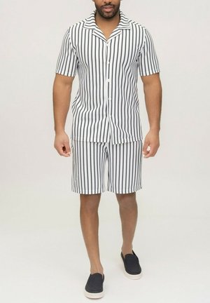 Homme portant une chemise blanche à manches courtes avec boutons et un short assorti à rayures noires verticales, associé à des chaussures noires sans lacets.