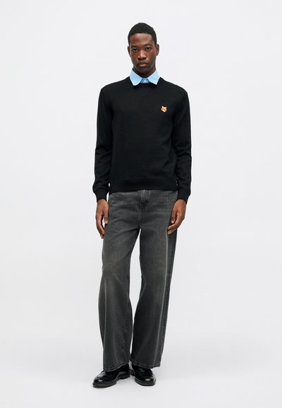 Ung mann iført en svart genser med en revelogo, lys blå skjorte med krage, vide mørke jeans og svarte dressko som står foran en hvit bakgrunn.