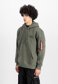 Sudadera verde oliva de tela suave, con un bolsillo tipo canguro, cordones ajustables y una etiqueta roja en la manga izquierda.