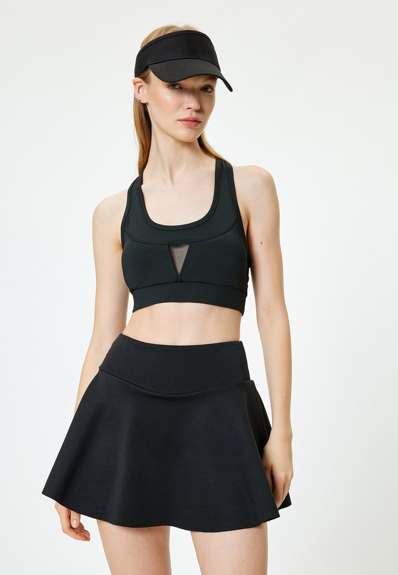 Svart atletisk outfit med en åtsittande sport-bh med nätdetaljer och en urtillverkad skort. Materialet ser slätt och stretchigt ut.