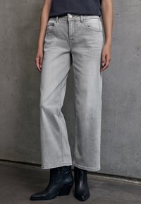 Persona che indossa jeans a gamba larga grigio chiaro e stivaletti neri in pelle, in piedi di fronte a uno sfondo di muro di cemento.