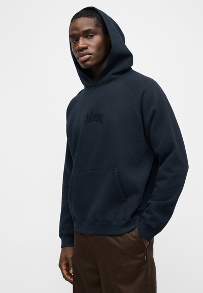 Νέος άνδρας φορώντας μπλε-γκρι φούτερ με κουκούλα και μπροστινή τσέπη και σκούρο καφέ παντελόνι, που στέκεται μπροστά σε απλό ανοιχτόφωτο φόντο.