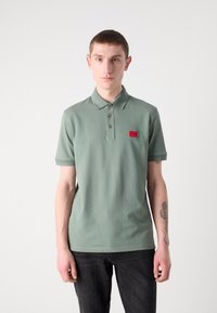 Lys grøn polo skjorte med knaplukket krave, korte ærmer og en rød mærke på venstre bryst. Bæres med mørke jeans.