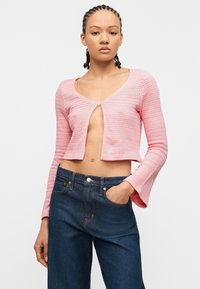 Νεαρή γυναίκα με πλεξουδάκια στα μαλλιά, φορώντας ένα κοντό ροζ πλεκτό ζακέτα με ένα κουμπί και σκούρα μπλε τζιν ψηλόμεσα, με το χέρι στην τσέπη.