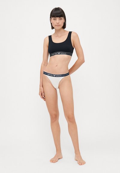 Soutien-gorge de sport noir avec une bande élastique blanche arborant le logo « Emporio Armani », associé à un string blanc. Tissu lisse, design minimaliste.