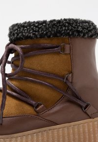 Bota de piel y ante marrón con cordones, que presenta un cuello de piel sintética negra, suela de plataforma texturizada y ojales de metal para los cordones.