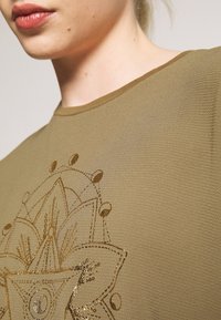 Gros plan sur une personne portant un haut texturé vert olive avec un motif floral mandala doré brillant sur la poitrine.