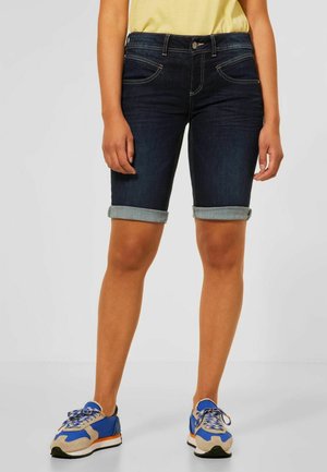 Persona con pantalones cortos vaqueros oscuros remangados, camiseta amarilla y zapatillas azules, beige y naranjas, de pie frente a un fondo liso.