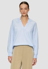 Chemisier rayé bleu clair avec un col volanté et des manches longues, fabriqué en tissu doux. Assorti à un pantalon beige.