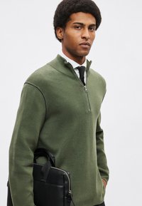 Πλεκτό πουλόβερ με φερμουάρ σε ελαιόχρωμο πράσινο με ψηλό γιακά, συνδυασμένο με λευκό πουκάμισο και μαύρη γραβάτα. Μαύρη δερμάτινη τσάντα κρατημένη στο αριστερό χέρι.