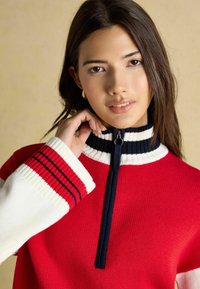 Roter Strickpullover mit hohem Kragen und Reißverschluss, der weiße und Marineblaue Streifenakzente am Kragen und an den Ärmeln aufweist.