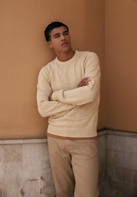 Jersey crema con un tejido texturizado, escote redondo y puños acanalados, superpuesto a una camisa de color óxido. Combinado con pantalones de un beige claro.
