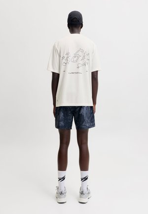 Homme portant un t-shirt blanc avec un motif graphique, un short noir à motifs, des chaussettes blanches, des baskets argentées et une casquette noire, debout de dos sur un fond uni.