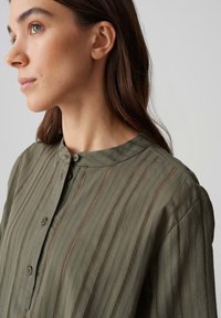 Blusa verde oliva con colletto rotondo, caratterizzata da strisce verticali e dettagli con bottoni lungo la parte anteriore. Tessuto leggero e strutturato.