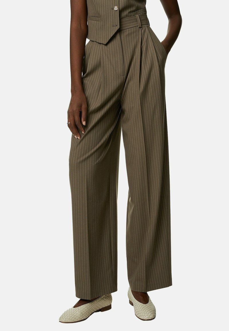 Femme portant un pantalon large taille haute à fines rayures marron, un gilet assorti et des chaussures plates blanches tissées, debout avec les mains dans les poches.