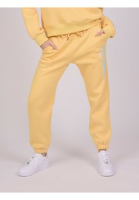 Gule fleece joggerbukser med elastiske cuffs, snøre i taljen, sidelommer og blå grafisk detalje på benet. Matcher med hvide sneakers.