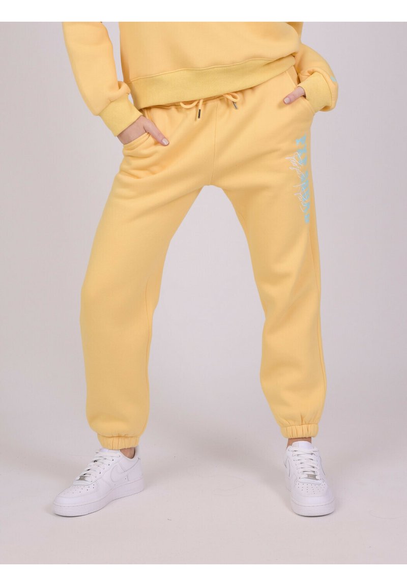 Gule fleece joggerbukser med elastiske cuffs, snøre i taljen, sidelommer og blå grafisk detalje på benet. Matcher med hvide sneakers.