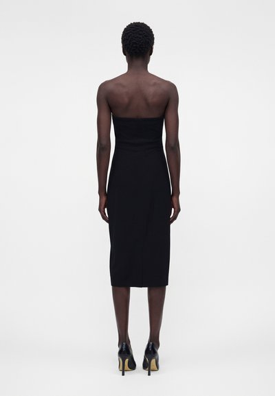 Γυναίκα με κοντά σγουρά μαλλιά που φοράει μαύρο μίντι φόρεμα χωρίς τιράντες και μαύρα ψηλοτάκουνα παπούτσια, στέκεται με την πλάτη προς τη κάμερα σε απλό φόντο.