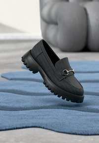 Mocassin en tissu noir à motif à fines rayures, bout rond, ornements argentés et semelle épaisse en caoutchouc noir à texture rainurée.