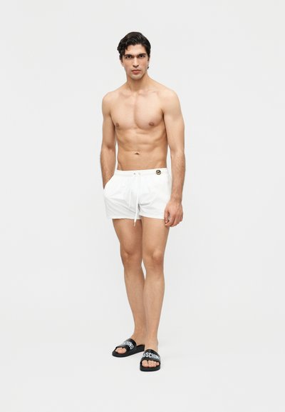 Άνδρας χωρίς μπλούζα, φορώντας λευκά μαγιό σορτς με λεπτομέρεια λογότυπου και μαύρα σανδάλια slide με λευκές επιγραφές, στέκεται μπροστά σε απλό φόντο.