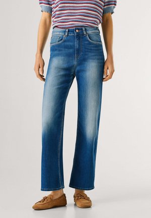 Persona che indossa jeans a vita alta di colore blu medio con gamba dritta, una maglia a maniche corte a righe e scarpe stile mocassino marroni.