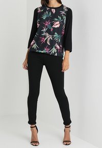 Musta kukkakuosiset puserot, joissa on 3/4-hihat, tropiikkikuosiset vaaleanpunaisena ja vihreänä, yhdistettyinä mustiiin joustaviin housuihin ja nauhakorkokenkiin.