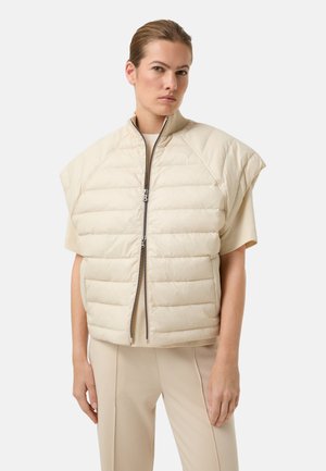 Kvinde iført beige quiltet ærmeløs vest med lynlås over kortærmet skjorte og matchende beige bukser mod en ensfarvet baggrund.