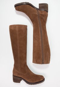 Botas de ante marrón hasta la rodilla con un acabado texturizado, cremallera lateral y tacón apilado. Presentan una punta redondeada y costuras decorativas.