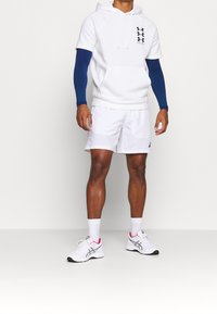 Atleta che indossa un hoodie bianco a maniche corte con maniche blu, pantaloncini sportivi bianchi, calzini bianchi e scarpe da corsa bianche con dettagli neri e rossi.