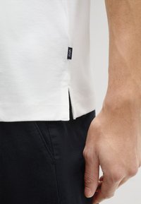 Witte katoenen t-shirt met zijsplitten, voorzien van een zwart label. Gekocht in combinatie met zwarte gestructureerde broek, waarbij een hand de tailleband vasthoudt.