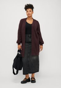 Cardigan lungo bordeaux, lavorazione a maglia testurizzata, davanti aperto, abbinato a una maglietta nera e una gonna di denim. Gli accessori includono una borsa nera e calzature.