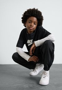 Melna Nike t-kreklā ar baltu logo, uzlikta virsū baltai garajai kreklai, kopā ar melniem džogiem un baltām sporta kurpēm, pozēta sēdus stāvoklī.