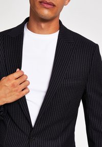 Skräddarsydd svart randig kavaj med en knapp, snedskurna slag och bröstficka, bärs över en vit t-shirt.