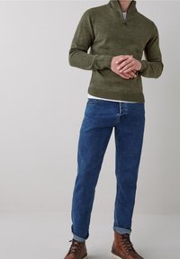 Pull en maille vert olive avec un col montant, associé à un jean bleu. Le mannequin porte des bottines marron à lacets. Texture lisse, coupe ajustée.