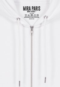 Felpa bianca in cotone con zip, caratterizzata da una zip argentata, coulisse bianche e un'etichetta con logo e istruzioni per la cura stampate all'interno.