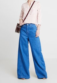 Donna che indossa una blusa a strisce rosa e bianche, jeans larghi blu, borsa a tracolla marrone e sandali, in piedi contro uno sfondo semplice.