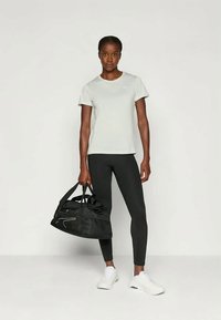 Šviesiai pilka sportinė marškinėliai, juodos leginsai ir baltos sportbačiai. Modelis laiko juodą sportinį krepšį su baltu akcentu. Paprastas dizainas, prigludusi forma.