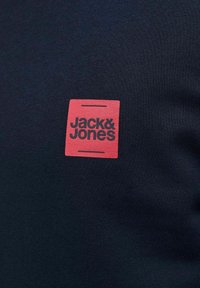 Rotes Quadrat-Logo mit schwarzem Text "Jack & Jones" auf dunklem marineblauen Stoffhintergrund.