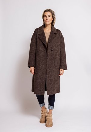 Woman standing wearing a long brown textured coat, rolled-up dark jeans, and beige fur-lined ankle boots against a plain light background.