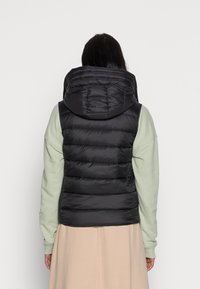 Schwarze Steppweste mit Kapuze, gestepptem Design und glänzendem Material. Über einem hellgrünen Langarmshirt getragen, von hinten präsentiert.