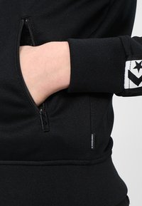 Black zip-up sweatshirt featuring a side pocket, ribbed cuffs, and a contrasting logo patch with star and chevron design.