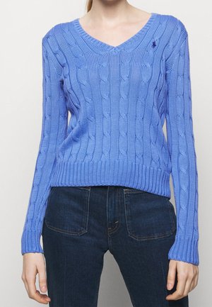 Femme portant un pull en maille torsadée bleu clair à col en V avec manches longues et un jean bleu foncé sur un fond uni.