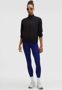 Kvinna i svart zip-up jacka, blå leggings, vita sneakers och strumpor, med solglasögon, som står framför en enkel ljus bakgrund.