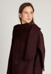 Bordeaux wollen jas met een brede kraag, een dubbelbreasted ontwerp met zwarte knopen en een verlengd gedrapeerd detail op de schouder.