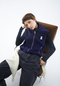 Marineblaue Fleece-Weste mit Reißverschluss, die ein weißes Emblem zeigt, kombiniert mit dunklen Jeans und einer cremefarbenen Strickschicht, die über den Oberschenkel drapiert ist.