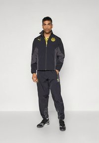 Man staat en draagt een zwarte en grijze Puma sportjas en broek met gele Borussia Dortmund-logo's op de jas en broek.