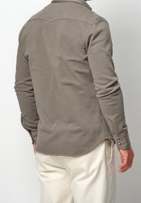 Chemise à manches longues grise en tissu doux, avec un ourlet arrondi, des poignets boutonnés et un col classique, portée avec un pantalon de couleur claire.