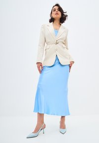 Beige kavaj med bälte över en ljusblå satin slipklänning, kombinerad med glittriga blå slingback-klackar. Släta texturer och skräddarsydd passform.