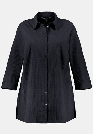 Chemise noire à manches longues, en tissu texturé, avec un col et des fentes sur les côtés pour plus de liberté de mouvement.