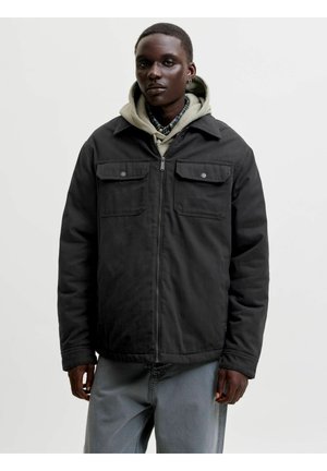 Jeune personne portant une veste noire zippée sur un sweat à capuche beige et une chemise à carreaux, debout devant un fond clair uni.